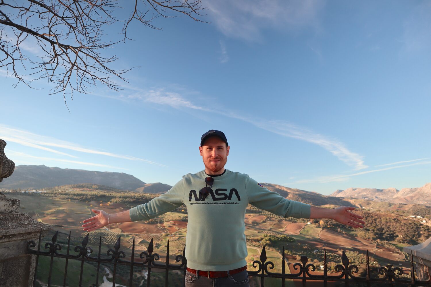 A man with outstretched arms stands in front of a scenic overlook with mountains and valleys in the backdrop.