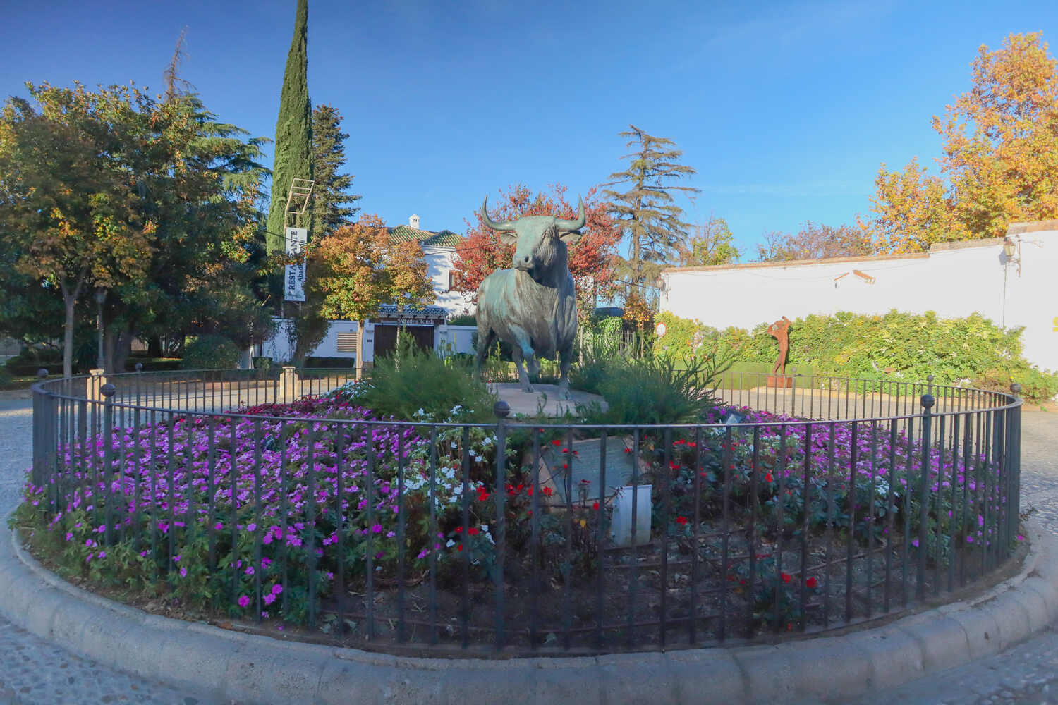 A vibrant flower garden with a central statue of a bull, surrounded by trees and a clear sky above.